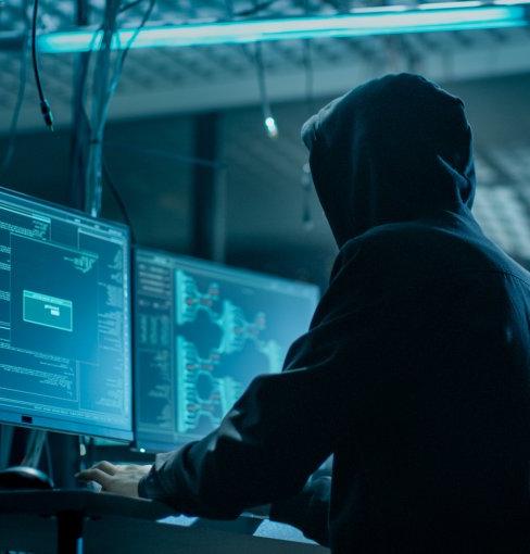 Shot from the Back to Hooded Hacker Breaking into Corporate Data Servers from His Underground Hideout. Place Has Dark Atmosphere, Multiple Displays, Cables Everywhere.